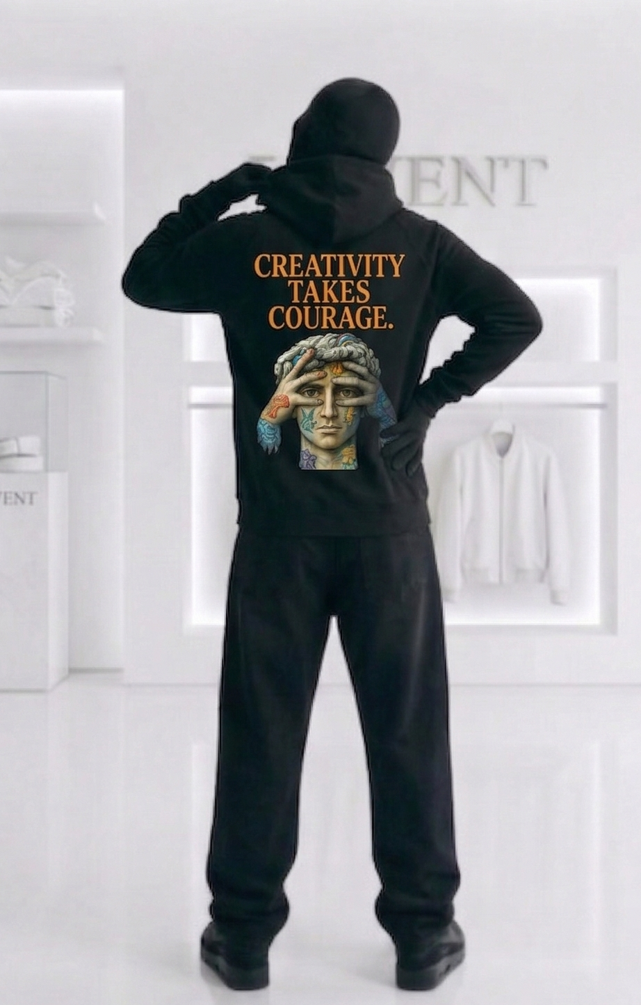 Person wearing a black hoodie with text and a gold graphic, standing in a white room.